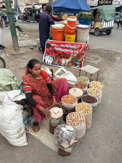 This is a photograph taken at street level, likely in a busy marketplace in Karachi, Pakistan. The scene depicts a woman in a vibrantly patterned red and orange outfit, with black sleeves and red bangles, sitting on the ground amidst sacks of produce and nuts. She appears to be a vendor, as she is surrounded by large transparent bags filled with various nuts and dried fruits, including what look like pistachios, almonds, and possibly dates. One hand is resting on a pile of what might be fresh dates or similar produce.
In the background, a multi-colored cart is visible, covered by a green tarp, with what seems to be a food or beverage stall signage in red. The signage, in Urdu script, advertises "Imran's Special 'Aloo Bukhara' and Lemon Water, and Rooh Afza," with a note about parcel arrangements. Several large insulated containers, likely for drinks, are stacked on the cart.
Other elements of the scene include a blue umbrella, some greenery, and the general urban environment of a marketplace with dirt and debris on the ground. In the mid-ground, two men are standing near the cart, and in the background, a small three-wheeled vehicle and motorcycles are visible, suggesting a bustling, active environment. The lighting suggests it is daytime. The woman's eyes are closed, and her expression is serene, possibly indicating rest or quiet concentration amidst the activity. The overall impression is of a vibrant street-side business operation in Pakistan.
