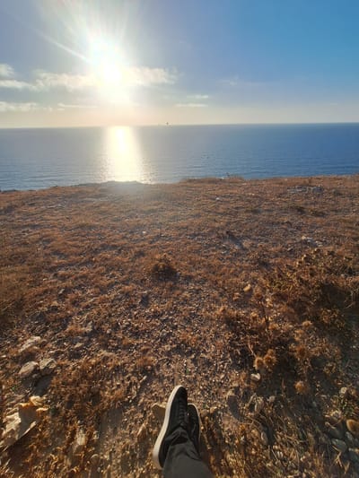 The image displays a person's feet wearing black sneakers and dark pants, standing on dry, brown vegetation and rocky terrain. The scene overlooks a vast body of water, likely the ocean, with a clear blue sky above. The sun is setting or rising, casting a bright, warm glow and creating a shimmering reflection on the water's surface. Wisps of clouds are visible in the sky, and a slight haze hangs over the horizon where the water meets the sky. The landscape appears arid, with sparse, dry plants and scattered stones. The time of day is clearly indicated by the low angle of the sun and the intense golden light. The location, Ahrara, Morocco, suggests a coastal setting with a potentially dry, Mediterranean climate. There is no visible text in the image. The overall mood conveyed is one of peaceful contemplation or perhaps the end of an adventurous day.
