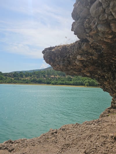 This media file depicts a scenic view of a body of turquoise water, likely a lake or reservoir, adjacent to a natural embankment. The foreground is dominated by a rocky overhang, offering a textured and earthy perspective. To the right of the water, a sandy and pebbly cliff face rises, with the water lapping at its base.
Beyond the water, a lush green landscape unfolds. A dense forest lines the shore, with trees of varying shades of green creating a rich tapestry. The terrain appears to be hilly or mountainous, with the forest covering the slopes. The sky is a clear, bright blue with scattered wispy clouds, suggesting a fair weather day. Sunlight glints off the surface of the water, creating a shimmering effect.
The image is oriented vertically, with the rocky overhang appearing at the top and the water filling the majority of the frame. The overall impression is one of natural beauty and tranquility. There are no people or human-made objects prominently visible, emphasizing the pristine, natural setting. The time of day appears to be daytime, given the bright sunlight and clear sky. The location cues, such as the type of vegetation and terrain, along with the provided context of Nanay, Uzbekistan, suggest a rural or natural park setting. No text is visible in the image.