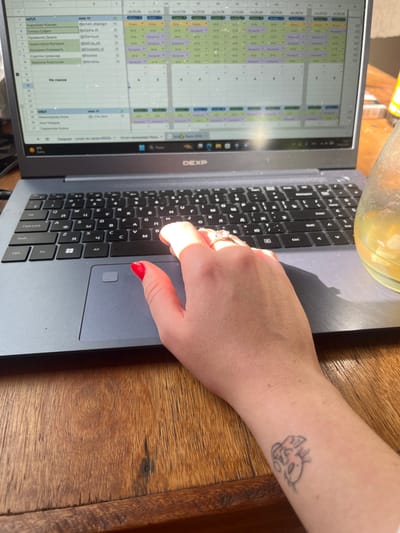 The image shows a close-up view of a person's left hand using a laptop on a wooden table. The laptop is open and displays a spreadsheet-like interface with names and dates. The person's hand, with red nail polish and a ring on the index finger, is positioned over the touchpad. To the right of the laptop, a glass with a yellow liquid is partially visible. The background shows a wooden surface, which is consistent with furniture found in a home or office environment. The image is taken from a top-down perspective, focusing on the interaction between the hand and the laptop. The scene is likely indoors, as indicated by the artificial lighting and the presence of a laptop. There are no discernible landmarks or contextual clues within the image that would allow for the specific identification of the city of Luque, Paraguay.