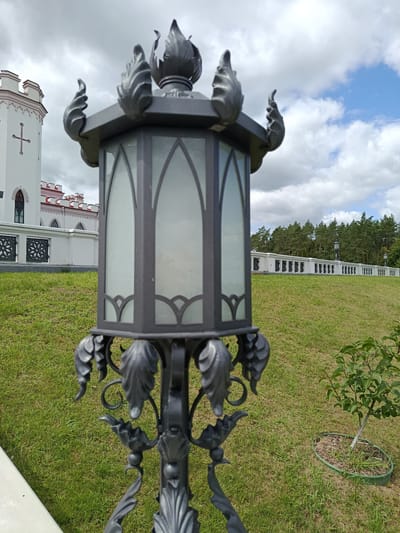 This is a medium shot, eye-level, outdoor photograph of a decorative lantern in the foreground, with the Kosava Palace (also known as Puslovsky Palace) in the background. The lantern is black and ornate, with glass panels and flame-like embellishments at the top. It is positioned on a grassy lawn. Behind the lantern, a white building with red trim and crenellations is visible. The building has arched windows and a cross emblem. A white fence with decorative black metal inserts runs horizontally across the middle ground. A line of trees forms the background, with a cloudy sky overhead. The overall scene depicts the grounds of the Kosava Palace in Kosava, Belarus. There are no people or animals in the image.