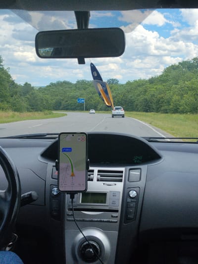 The image is a first-person perspective from inside a car, looking forward through the windshield. The car's dashboard is visible, with a smartphone mounted on a holder displaying a navigation app with a route and distance of 500 meters. The car is on a road, with green trees and foliage lining both sides. In the distance, another white car is visible on the road, which curves to the left. A blue road sign is also present in the distance. The sky is cloudy with patches of blue. The scene appears to be a rural or suburban setting. The car's interior shows a steering wheel on the left, the dashboard with various controls and a digital clock displaying 12:40, and a passenger air vent. The overall lighting suggests it is daytime. Based on the available visual information and its distinct road and landscape features, this scene is likely located in the vicinity of Vadul lui Voda, Moldova, a town known for its scenic routes and natural surroundings.