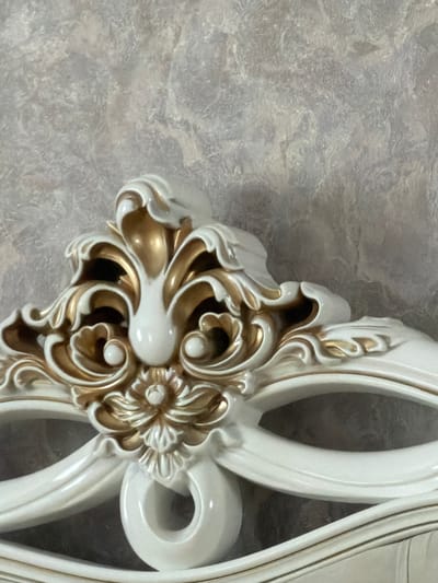 This is a close-up, eye-level, medium shot of an ornate decorative element, likely a furniture crest or architectural molding, rendered in white with gold accents. The material appears to be a resin or carved wood with a glossy finish. The design is elaborate, featuring acanthus leaf motifs, scrolled embellishments, and a central floral element. The background is a textured wall, possibly wallpaper or plaster, in muted shades of grey and beige. The lighting is soft, casting subtle shadows that highlight the depth and detail of the decorative piece. The image is devoid of any people, animals, or signs of activity. The precise location in Karshyga-Aul, Russia cannot be determined from the visual information provided.