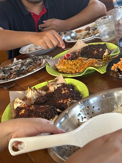 The image depicts a close-up view of a table laden with food, likely in a restaurant or home setting in the city of A R A, Indonesia. Several plates of food are visible, including what appears to be grilled fish or lobster with a dark sauce, and another dish with a bright yellow topping. There are also plates with rice and what seems to be clams or shellfish. A glass of water with ice is present, along with cutlery and a serving spoon. Two individuals are partially visible in the frame. One person, wearing a dark blue polo shirt with a red collar, has their arm extended over a plate of food. Another person's hands are visible in the foreground, holding a white serving utensil over a bowl of rice. The overall impression is a casual meal, possibly a gathering or a family dinner, with a focus on seafood dishes. The wooden table surface suggests a rustic or traditional dining environment.