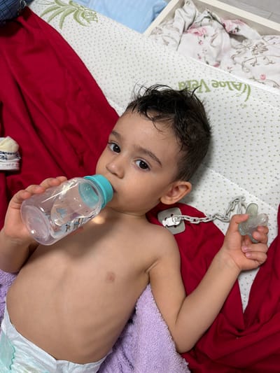 A young child, approximately 2-3 years old, lies on a bed. The child is partially covered by a red blanket and a lavender towel, wearing a diaper, holding a clear plastic bottle with a light blue lid in their right hand and a pacifier with a gray plastic chain in their left hand. The bedding consists of a white, patterned mattress with text visible as "Aloe Vera" and a decorative, floral-patterned item in a white tray on the left. The child's gaze is directed towards the camera. The location is unknown, and there is no indication it is Iasenovets, Bulgaria.