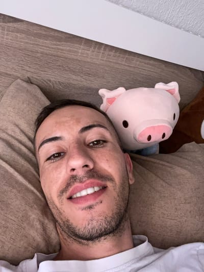 A first-person perspective selfie shows a man lying down next to a stuffed pig. The man is looking at the camera with a slight smile, his mouth open to reveal white teeth. His skin is tanned, and he has dark hair and eyebrows. He is wearing a white t-shirt. The stuffed pig is pink with black eyes and pink ears. The background appears to be a bed or sofa with a wooden surface on the left. The lighting suggests it might be daytime, and there are no strong indications of the weather or specific location beyond what's implied by the indoor setting. The man's expression is relaxed and friendly.