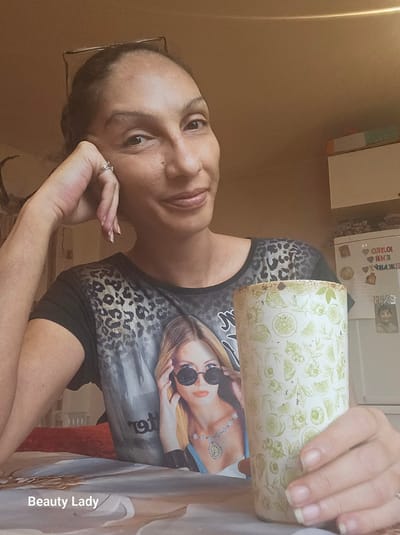 A medium shot shows a woman holding a floral-patterned cup. She is wearing a black t-shirt with a graphic of a woman wearing sunglasses and a leopard print overlay. She has dark hair, light skin, and is wearing a ring. She appears to be indoors. The background is a beige wall. There is a sign in Cyrillic above her. There is a watermark "Beauty Lady" in the lower right. There is no information available to determine the specific location as Fredrikstad, Norway.