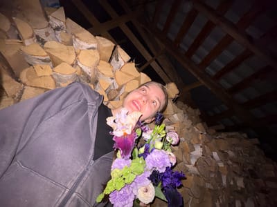 The image shows a young woman lying down, holding a bouquet of flowers. She is wearing a grey zip-up hoodie over a dark shirt. Her head is tilted back, and she is looking upwards with a smile. The background consists of a large pile of chopped firewood stacked against a wall, and overhead, a wooden roof structure is visible. The scene appears to be in a dimly lit storage area, possibly a shed or barn, located in Нижние Муллы, Russia. The woman is positioned in the foreground, with the firewood and roof structure forming the backdrop. The bouquet contains a variety of flowers in shades of purple, pink, and green. The lighting suggests it is nighttime or a very dark indoor environment.