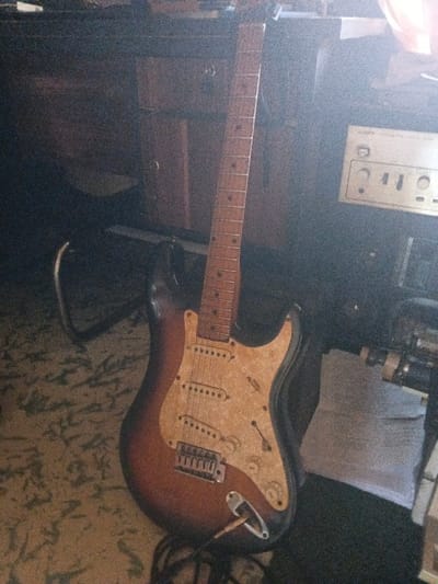 An indoor scene in Dogsenang Gorphoog, Bhutan, depicts one electric guitar standing upright. The guitar features a three-tone sunburst body (brown, dark brown, black), a cream-colored pickguard with a decorative, swirling pattern, three visible single-coil pickups, a tremolo bridge, and three control knobs. A cable is connected to the output jack. The fretboard is a light-toned wood with dark circular fret markers. In the background, to the left of the guitar, a dark wooden cabinet with two visible drawers and round pulls is present. The floor is covered with a patterned carpet displaying light and dark organic shapes. To the right of the guitar, a silver-toned audio receiver or amplifier, marked 'SONY' and featuring multiple controls, is visible. This area is subject to significant glare from a strong light source located upper-right. Below the receiver, dark equipment and a crumpled white sheet of paper are discernible. No individuals are present. The image exhibits a grainy texture.