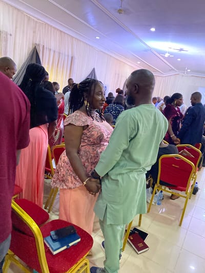 A gathering of people is taking place indoors. The setting is likely a church, suggested by the presence of a Holy Bible and the prayerful posture of some attendees. The location could possibly be Miango, Nigeria, based on information provided in the prompt. A man in a light green outfit is in the foreground, with his hands clasped. Next to him is a woman wearing a dress with pink floral patterns. Other people are seated in red chairs. Some of them are praying or watching someone. The environment is indoors.