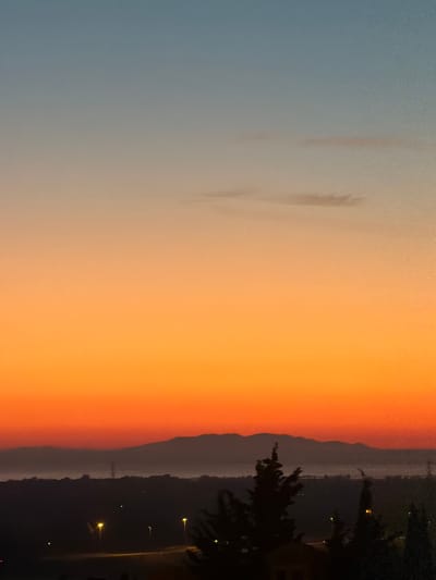 The image captures a sunset view from Tarquinia, Italy, displaying a gradient sky transitioning from blue to orange. A mountain range silhouettes against the vibrant sky. Streetlights are visible along a road at the base of the mountains, and trees are silhouetted in the foreground.