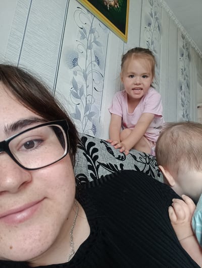 A selfie taken indoors, likely in a residential setting in Ust-Tuntor, Russia, shows three people: a woman in the foreground wearing glasses and a black top, a young girl sitting on a patterned sofa in the middle ground wearing a pink t-shirt, and a baby whose head and shoulders are visible on the right side of the image. The background features striped wallpaper with a floral pattern and a framed picture on the wall. The lighting appears to be natural, coming from an unseen source, creating soft shadows. The overall scene suggests a casual, domestic moment.