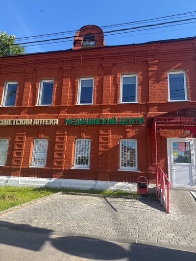 In the city of Vereya, Russia, a two-story red brick building with arched windows on the upper floor is shown. The building has several rectangular windows on both floors, some with decorative brickwork above them. A sign in white and green Cyrillic lettering spans the upper facade, reading "СОВЕТСКАЯ АПТЕКА - МЕДИЦИНСКИЙ ЦЕНТР" (Soviet Pharmacy - Medical Center). A red awning extends over the entrance to the building, which is a white door with a glass panel. A ramp leads to the entrance, flanked by a red railing. A red trash receptacle is situated near the entrance. The ground in front of the building is paved with grey bricks, transitioning to asphalt in the foreground. Green grass grows along the base of the building. The sky is clear and blue, with a few dark power lines running across the top of the frame.