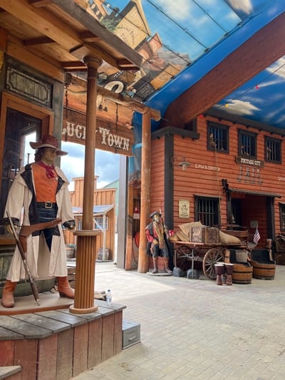 The image depicts an indoor Western-themed attraction, likely in the city of Collendoorn, Netherlands. In the foreground, a mannequin dressed as a cowboy stands on a wooden platform, holding a rifle. The mannequin wears a brown hat, an orange vest, a long cream-colored coat, and brown boots, with a gun belt and holster. Behind the cowboy mannequin, a sign reads "LUCKY TOWN".

To the right, a wooden building facade mimics a Wild West jail. It is painted red and features barred windows. A sign above the entrance reads "POSTAGE CITY JAIL". A wagon laden with sacks and crates is parked in front of the jail building, alongside several wooden barrels and two drums. Another mannequin dressed as a cowboy stands near the wagon, holding a rifle.

The ceiling is decorated to resemble a blue sky with clouds and painted buildings, giving the impression of an outdoor scene within an enclosed space. The ground is paved with cobblestones. The overall scene is designed to evoke a historical Western town setting.