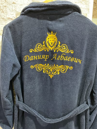 The image shows the back of a dark blue terrycloth robe. Embroidered on the back of the robe in gold thread is a crest featuring a lion's head wearing a crown, surrounded by decorative scrolls. Below the crest, the name "Данияр Агбаевич" is embroidered in elegant script. The robe is tied with a sash at the waist. The background appears to be a textured wall, possibly a brick or stone pattern, with a light colored wall to the right. The scene is indoors. There are no people or animals visible in the image. The robe is likely a personal item, possibly a gift or a custom-made piece. The embroidery suggests a personal touch, perhaps for a special occasion or as a sign of luxury. The location is specified as Бескөл ауылдық округі, Kazakhstan.