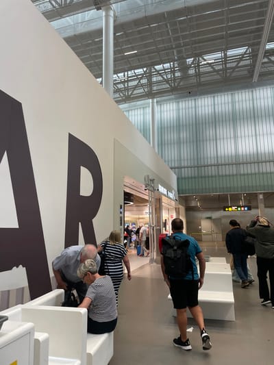 The image shows a view of an airport terminal in Granadilla, Spain (28.047, -16.58). The scene is dominated by a large white wall with a brown "R" printed on it. There are four people visible in the image. A man wearing a blue shirt and black shorts is walking towards the right, carrying a black backpack.  Behind him is a woman wearing a striped top and dark pants, walking towards the left.  A second woman, also wearing a striped top, sits on a white bench with a man in a grey shirt and blue jeans. The airport terminal features a large, white ceiling with exposed metal beams. The wall behind the "R" has vertical, translucent green panels. There is a shop with a black door labeled "TRAVEL PRODUCTS" to the right of the "R," and a large window behind the shop, showing an open area of the airport. Two white benches are placed next to each other.  Two men, both wearing dark shirts, walk towards the left, one carrying a large bag over his shoulder, and the other with a smaller black bag.  The man with the large bag also appears to be talking on his phone.  All of the individuals are facing the right.