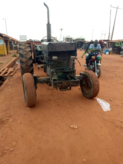 The image depicts a vibrant street scene in Mbar, Nigeria, dominated by an old, front-facing tractor positioned on a reddish-brown dirt road. The tractor appears weathered and robust, featuring a prominent vertical exhaust pipe, large, deeply-treaded rear tires, and smaller front wheels. Its metal body is rusted and shows signs of extensive use, with dirt covering its frame and components.
To the right of the tractor, a man is seated on a red motorcycle, looking towards the camera. He is dressed in a light-colored, possibly patterned shirt and dark trousers. The motorcycle has a round headlight and a distinctive circular emblem on its front, which displays vertical stripes of green, orange, and green. In the background, other individuals and motorcycles are visible, indicating ongoing street activity.
The setting is characterized by a wide, unpaved road composed of reddish-brown laterite soil, typical of the region. Simple, rustic structures, possibly market stalls or shops with corrugated iron roofs, line both sides of the road. Utility poles and power lines are visible against a bright, possibly overcast, daytime sky. Scattered debris, including a white plastic bag, lies on the ground. No legible text is clearly visible on any signs or structures within the frame. The overall atmosphere suggests a bustling, everyday scene in a semi-urban or rural Nigerian locality.
