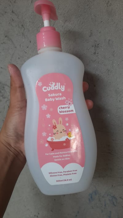 The image shows a person's hand holding a pink and white pump bottle of "Cuddly Sakura Baby Wash" with "cherry blossom" scent. The bottle has a pink label with illustrations of a bunny in a bathtub and flowers. The text on the label indicates that the product is "For Calm and Moisturised Skin," "Made for Babies," and "Gentle on Skin." It also states "Silicone Free, Paraben Free, Gluten Free, Phalate Free" and has a volume of "500ml (16.9 oz)". The background is a textured grey wall. The image was taken in Kawit, Philippines.