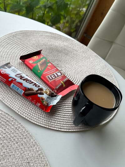 A top-down shot captures a breakfast scene with two chocolate bars and a mug of coffee. The setting appears to be indoors, possibly near a window given the glimpses of greenery outside. The light suggests it's daytime.
Dominating the frame are two chocolate bars placed on a woven placemat. The bar on the left is red and features Russian text that includes "СТЕП" and "ШОКОЛАД С ОРЕХАМИ" (Chocolate with Nuts). It also mentions "ЛЕСНОЙ ОРЕХ, АРАХИС И КАРАМЕЛЬ" (Hazelnut, Peanut, and Caramel), with an image of a chocolate bar with nuts and caramel.
To the right, and slightly open, is a Kinder Bueno bar. Its distinctive white and red packaging is visible, with the "bueno" logo prominently displayed. The bar itself shows its wafer and chocolate construction, along with whole hazelnuts.
A black mug, filled with a light brown beverage, likely coffee with milk, is positioned above the chocolates. The mug has a sleek, modern design with a thick handle. The placemat under the items is a circular, textured, light-colored weave. The overall arrangement on a plain white surface suggests a simple, everyday moment of enjoying a sweet treat with coffee. The presence of "EAC" on the Kinder Bueno packaging indicates compliance with Eurasian Customs Union standards.