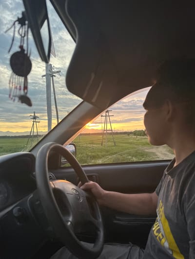 An interior shot from the driver's perspective inside a car shows a person's head and shoulders from the side. The person is looking out the side window towards a landscape with a sunset. In the foreground, a steering wheel and part of the dashboard are visible. The environment outside the car appears to be a rural setting with fields, power lines, and mountains in the distance. The lighting suggests it is either sunrise or sunset. The image is set in the village of Ketkino, Russia.