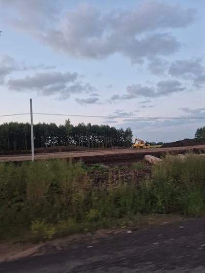 In Kabanikha, Russia, a yellow excavator is operating on a dirt road. The excavator is in the middle distance, to the right of the frame. The background consists of a dense line of trees, and the sky above is partly cloudy. In the foreground, there is thick green vegetation. The overall scene depicts a construction or land clearing site.