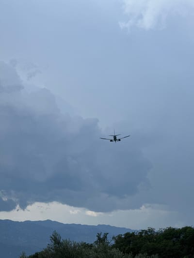 An airplane is seen flying against a cloudy sky, possibly approaching an airport near Pelinovo, Montenegro. The airplane is positioned in the upper right quadrant of the frame, appearing as a dark silhouette against the lighter sky. Below the sky, a range of mountains is visible, their peaks softened by distance and the atmospheric conditions. The foreground is occupied by dense green trees and foliage, suggesting a natural landscape. The overall lighting suggests an overcast or possibly approaching storm, contributing to a muted color palette. The scene is devoid of human figures.