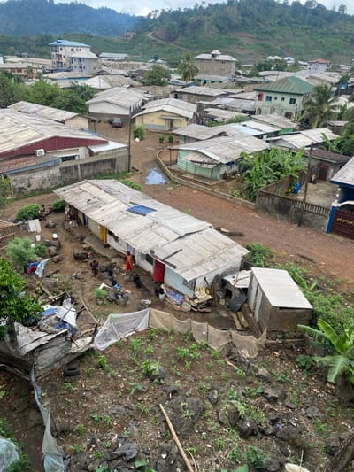 The image shows a scene from a residential area in Bonadikombo, Cameroon, located at the coordinates 4.059 latitude and 9.231 longitude. The scene is dominated by a collection of simple, single-story homes with corrugated metal roofs. The houses are situated on a hillside, with some vegetation growing around them. Several individuals are visible in the scene, most of whom are adults, engaged in daily activities. There are also several objects present in the scene, including a few cars, a variety of plants and trees, and other detritus. The overall scene is one of a typical residential neighborhood in a developing country.