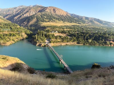 An aerial, wide shot captures a serene natural landscape with a prominent bridge spanning a tranquil, turquoise lake. The lake's surface is rippled, indicating gentle movement of the water. The bridge, a long, slender structure supported by concrete piers, is the focal point of the scene, extending from one shore to the other. A small boat is visible in the lake, leaving a white wake behind it.
The surrounding environment is characterized by steep, mountainous terrain covered in lush green vegetation, predominantly trees and scattered settlements. The mountains exhibit rough, textured surfaces, with visible geological formations and shadows cast by the sunlight, suggesting a time of day early morning or late afternoon. The sky above is a clear, pale blue, devoid of clouds.
The foreground of the image features a dry, grassy hillside with a scattering of scrubby bushes, adding texture and depth to the composition. The overall impression is one of a remote, picturesque location where nature dominates the landscape. While the prompt mentions Nanay, Uzbekistan, the visual information itself does not provide explicit clues to confirm this specific location, relying solely on the visual elements present.