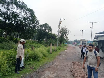 The image depicts a scene in Benapole, Bangladesh, featuring a dirt road with some overgrown vegetation on the left and industrial-looking structures on the right. Several people are visible. In the foreground, two men are standing on the grassy verge. The man on the left wears a light-colored patterned shirt, dark jeans, and a light-colored cap. The man next to him is partially visible and wears a dark shirt. Further down the road, a man in a light-colored t-shirt with "Original" printed on it, blue jeans, and a backpack is looking towards the camera and smiling. He is holding a phone in his right hand. Behind him, another man in a white shirt is taking a photo with a camera, and a man in a red shirt stands near a building. The road itself is unpaved and appears to have some potholes and a patch of exposed bricks. Overhead, utility poles with streetlights and electrical wires are present. The sky is overcast.