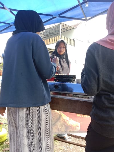 Three women are seen under a blue awning, two are partly visible from the side. The woman in the center, wearing a hijab, appears to be cooking on a small stovetop on a stand. She is using a utensil, likely a stick or skewer. The woman on the left wears a hijab, a blue sweater, and a long patterned skirt. A yellow propane tank is visible at the bottom left. A building is visible in the background.