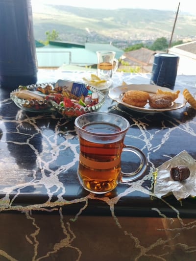 A high-angle medium shot captures a table set with tea and snacks, overlooking a city and mountainous landscape. In the foreground, a glass mug filled with amber tea sits on a dark, glossy table with a marbled pattern. A small, unwrapped chocolate lies to the right of the mug on a crinkled silver wrapper. Behind the tea, a glass dish overflows with assorted candies, and a plate holds three cookies. Two blue containers, one tall and dark, the other shorter and lighter, are also visible on the table. In the background, a picturesque view unfolds with a city nestled in a valley between rolling hills, suggesting a location like Darvag, Russia. The overall scene evokes a sense of leisurely enjoyment with a scenic backdrop.