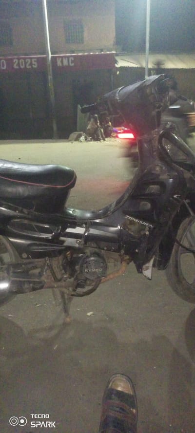 At night, a black Kymco Zongshen motorcycle parked on the street. The bike is the focal point, positioned towards the right side of the frame. Behind the bike, blurred figures are visible near a building with "KMC" and "O 2025" written on it. In the foreground, a single foot wearing a shoe appears on the right side of the image. The lower left corner features a "TECNO SPARK" watermark. Streetlights illuminate the scene.