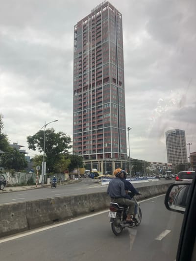 The image presents an urban street scene in An Hải Ward, Vietnam, viewed from a vehicle on an overcast day. A prominent, slender high-rise building stands in the upper center-right, characterized by a dark grey facade with a distinctive red-orange grid pattern of vertical and horizontal accents across its numerous stories and balconies. The word "PREMERA" is partially visible at its summit.

In the mid-ground, a multi-lane road is visible, partially separated by a low concrete median barrier. Two individuals are riding a dark-colored scooter in the lane adjacent to this barrier, moving away from the viewer. The driver wears an orange helmet, a light-colored plaid shirt, and light trousers. The passenger is clad in a dark blue top and dark trousers. The scooter's license plate displays "43C4 - 6877." Further down the road on the left, another individual on a scooter, wearing a blue shirt and white helmet, is observed.

Vegetation, including trees and shrubs, lines the left side of the road. Another tall, grey, multi-story building with a grid-like appearance is visible in the distant background to the right of the main high-rise. Various other vehicles, including cars, trucks, and yellow taxis, are present on the road further ahead. A street light pole is visible on the left. A wall with graffiti is present on the far left of the frame. The immediate foreground shows a portion of a vehicle's interior, including a side mirror on the right.