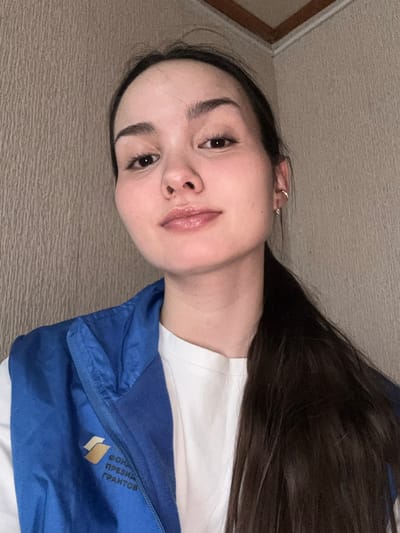 In an interior setting in Bayronovka, Russia, a young adult female with dark hair and a light complexion faces the camera directly. Her dark hair is pulled back, with a long section visible over her right shoulder. She wears a small gold hoop earring in her visible left ear. Her lips are glossy, and her expression is neutral with a slight smile. She is dressed in a white t-shirt layered under a royal blue sleeveless zippered vest. On the left chest of the vest, a light-colored logo is visible, bearing partial Cyrillic text that appears to read "ФОНД ПРЕЗИДЕНТСКИХ ГРАНТОВ" (Fund of Presidential Grants). The background features a textured, light brown wall, with a ceiling corner visible in the upper right quadrant of the image. The lighting is even and consistent with indoor conditions.
