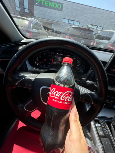 The image shows the interior of a car, viewed from the driver's perspective. A hand is holding a bottle of Coca-Cola in front of the steering wheel. Through the car's windshield, a parking lot is visible, with several cars parked and a building in the background. The building has a green sign with the word "TENDO" on it. The license plate of one of the visible cars is blurred. The car's dashboard and steering wheel are clearly visible. The image appears to have been taken in the city of Sarny, Ukraine. The presence of the "TENDO" sign on the building suggests it may be a store or shopping center in that city.