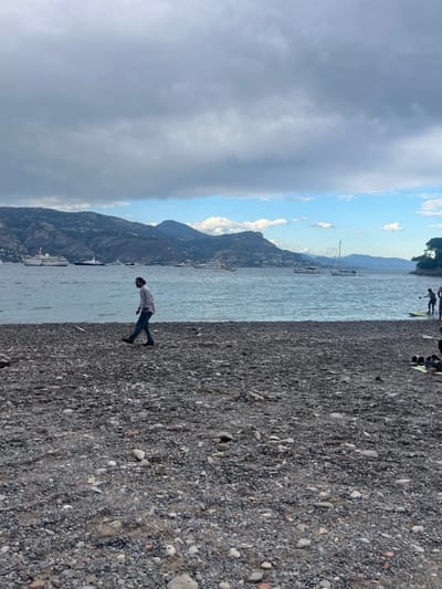 The image displays a wide view of a pebble beach and a bay in Saint-Jean-Cap-Ferrat, France, under a sky largely covered by dark gray clouds, with a segment of blue sky and white clouds visible above the distant horizon. The foreground consists of a coarse pebble beach, predominantly dark gray and brown. A male individual, exhibiting dark hair and a beard, dressed in a light-colored long-sleeved shirt, blue jeans, and dark shoes, walks from left to right along the water's edge. The calm, light blue-green water of the bay extends from the immediate shoreline to a distant, mountainous coastline. Several vessels are present on the water: multiple large white yachts are anchored in the mid-distance, and smaller boats, including a white sailboat, are positioned closer to the right. In the far right background, two individuals are partially visible near the water; one is standing on a yellow paddleboard holding a paddle, while the other is partly obscured. Further right on the beach, a person is seated on a light-colored mat, with dark objects positioned nearby.