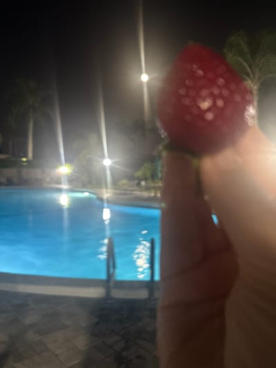 The image shows a close-up of a strawberry being held by a person's hand. The strawberry is red and has visible seeds. In the background, there is a blurred image of a swimming pool at night, with lights reflecting on the water. Palm trees are also visible in the distance, suggesting a tropical or subtropical location. The overall scene appears to be outdoors, possibly at a resort or hotel in Delray Beach, United States, given the presence of palm trees and a swimming pool at night.