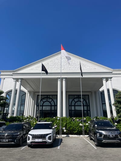 The image displays the exterior of a large, white building with neoclassical architectural features, located in Kayuambon, Indonesia. The building features a prominent triangular pediment adorned with intricate white bas-relief ornamentation, supported by several fluted white columns. Large arched windows or doors with black frames are visible on the facade.

Three flags are displayed on flagpoles in front of the building. The central flag is a red and white horizontal bicolour, positioned slightly higher than the two flanking flags. To its left and right are solid black flags.

In the foreground, a paved parking lot with interlocking brick-like pavers accommodates three parked cars. From left to right:
1.  A black Toyota Veloz, with license plate F 1042 AAO, is parked facing the building.
2.  A white Hyundai Creta, with license plate D 1598 AJH, is parked facing the building.
3.  A black Mitsubishi Pajero Sport, with license plate B 2502 SJG, is parked facing the building.
All cars are positioned within marked parking spaces.

A line of dense green bushes interspersed with white and light purple flowers runs along the base of the building, separating the parking area from the structure. Partially obscured within these bushes are dark-colored decorative statues resembling deer or similar animals. The sky is clear and deep blue, indicating bright, sunny weather.