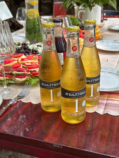 The image depicts an outdoor gathering on a wooden table, likely in Буран Снт, Russia, as indicated by the context. Three bottles of "Baltika 1" light lager are prominently displayed in the foreground. The bottles are golden yellow, filled with a pale yellow liquid, and have yellow caps with red and white labels. The labels feature the brand name "Балтика," the number "1," and text that reads "лёгкий лагер" (light lager).
In the background, a platter of food is visible, featuring circular slices of food topped with red tomato slices and green herbs, possibly appetizers or small dishes. There are also glasses on the table, including a wine glass on the left and a tall glass with what appears to be water and mint. Other elements in the background include a white pitcher or jug, some greenery from a plant, and parts of plates. The overall setting suggests a relaxed outdoor meal or social event. The lighting indicates it is daytime, with natural light illuminating the scene.