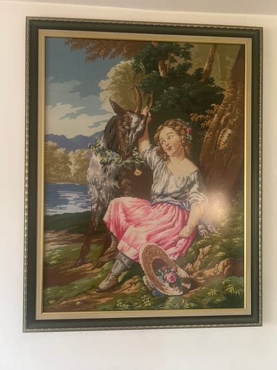 The image shows a framed needlepoint tapestry depicting a pastoral scene. In the foreground, a young woman with brown hair, wearing a light-colored off-the-shoulder blouse and a pink skirt, sits on the ground next to a tree. She is leaning on a goat or similar animal, which is adorned with flowers. The woman holds her hand up near the animal's head. A straw hat with flowers is placed beside her. The background includes a body of water, trees, and mountains. The tapestry is framed with a dark green and gold border. The tapestry is hanging on a white wall. The scene is not specifically identifiable as being located in Guliya, Bulgaria.