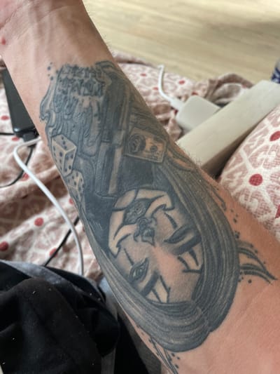 The image shows a close-up view of a tattoo on a person's forearm. The tattoo appears to be in a dark ink, possibly black and grey, and depicts a stylized face with flowing hair, dice, and what looks like money. The skin around the tattoo has some light hair. The background is blurred, showing a patterned fabric, likely bedding or upholstery, in shades of pink and white with red circular patterns. There is also a white cable and a dark object, possibly a phone charger or electronic device, visible in the background. The setting does not provide any information to place it in Paczkowo, Poland.