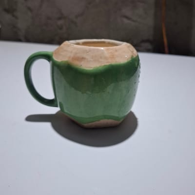A ceramic mug shaped like a coconut is resting on a white surface. The mug is primarily green with a tan-colored rim. A green handle is attached to the left side of the mug. The mug casts a shadow to its right. The background is out of focus and appears to be a textured wall. There is no visible indication of the city of Reudeuep, Indonesia, in this image.
