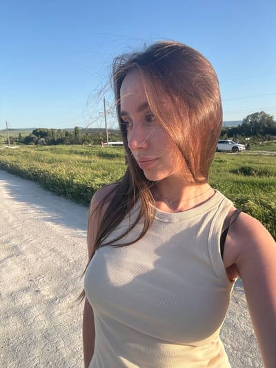 A young woman stands on a light-colored dirt path in Вишенское сельское поселение, Ukraine. She wears a light-colored ribbed tank top and a delicate necklace. Her hair is long and brown. A grassy field stretches out beside the path, and in the distance, there is a parked white vehicle. Overhead utility lines stretch across the clear blue sky.