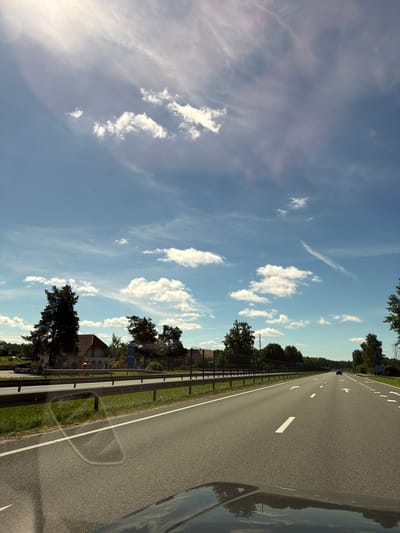 The image depicts a highway scene in Medemciems, Latvia. The perspective is from inside a moving vehicle, looking forward and slightly to the right. The road is asphalt with clear white lane markings, including arrows indicating direction. A metal guardrail runs along the side of the road, separating it from a grassy verge and then a line of trees. Beyond the trees, several houses with red-tiled roofs are visible, along with more greenery. A gas station with blue signage is also present.
The sky is a clear blue with scattered white cumulus clouds and some wispy cirrus clouds. The sunlight suggests it is daytime, likely late afternoon given the angle of the sun and the shadows. The overall atmosphere is calm and serene, conveying a sense of travel. The reflection on the car's surface shows a mirror image of the sky and trees. There is no visible activity of people. The blue sign at the gas station appears to have "00-24" printed on it, indicating 24-hour service.