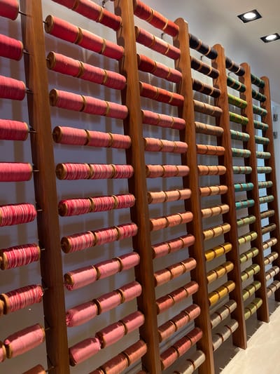 The image depicts a meticulously organized display of spools of thread arranged vertically on wooden racks. The spools vary in color, featuring shades of pink, red, brown, green, yellow, and white, creating a vibrant visual spectrum. The wooden racks are positioned against a plain, light-colored wall, and the lighting suggests an indoor setting. There are no people visible in the image. The overall impression is one of a textile shop or a craft store, possibly a boutique specializing in yarns or threads. The scene is static and orderly, emphasizing the craftsmanship and variety of the materials on display. The location is specified as Port De Pollença, Spain.