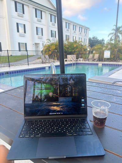 The image shows a laptop open on a wet, dark brown wooden table next to a swimming pool. The laptop screen displays what appears to be a poker game interface with multiple tables visible. Behind the table, a swimming pool with blue water and lounge chairs can be seen. In the background, a white hotel building with multiple windows and balconies is present, along with palm trees. A "NO DIVING" sign is visible near the pool fence. The environment suggests a resort or hotel setting, likely in a warm climate. The time appears to be late afternoon or early evening, given the lighting. There is a plastic cup with a dark beverage and ice cubes next to the laptop. The overall scene depicts someone working or playing poker outdoors by a pool.