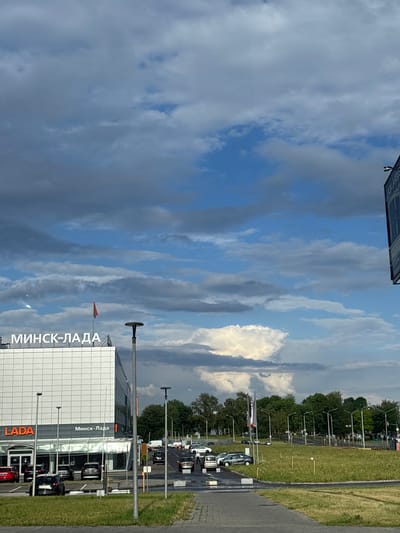 This is a vertical, eye-level, outdoor shot of a car dealership and its surroundings under a dynamic sky. The dealership, identified by "МИНСК-ЛАДА" signage in large white letters on a white, modern building, is prominent on the left. A red and green flag flies from a pole on the roof of the dealership. Below the large signage, smaller "LADA" lettering is visible in orange on the lower portion of the building, with "Минск-Лада" also appearing in white. The building has a glass-fronted showroom area. Parked cars, including a red Lada and several black cars, are visible in the foreground and in the dealership's lot.
The environment includes a paved area in front of the dealership, a grassy field, and a line of trees in the background. A large, cumulonimbus cloud formation dominates the sky, with streaks of dark clouds interspersed with patches of blue sky and brighter, lighter clouds. Streetlights and traffic signals are visible throughout the scene, indicating a road or intersection in the distance. Several cars are driving or parked on a road to the right of the dealership. A person can be seen near the parked cars in front of the dealership, and other figures are present further down the road. The overall scene suggests an overcast but potentially bright day in a suburban or semi-urban area.