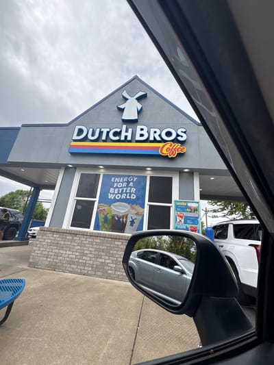 The image depicts a Dutch Bros Coffee drive-thru located in Tigard, United States, viewed from the interior of a vehicle. The building features a gray facade, blue trim, and a gabled roof displaying a white windmill logo. Prominent "DUTCH BROS Coffee" signage with a red, yellow, and blue stripe is visible on the upper facade. The lower portion of the building is faced with light-colored brick. A large window poster states "ENERGY FOR A BETTER WORLD" above images of three beverages, while an adjacent vertical sign displays menu items and prices.
The scene is set under an overcast sky. In the drive-thru lane to the left, a dark-colored SUV is stationary, with a person standing near its driver's side. Further along the lane to the right, a white SUV is also visible. The foreground includes the dark frame of the viewer's vehicle's A-pillar on the right and a significant portion of its passenger-side mirror, which reflects a silver sedan, likely positioned behind the viewer's vehicle in the queue. A person's head is discernible through the driver's side window of the reflected silver sedan. A blue metal picnic table or bench is partially visible on the left side of the concrete drive-thru pavement.