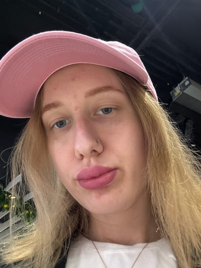The image displays a close-up, portrait-style view of a young person, likely female, looking directly at the camera. She has fair skin, light blue eyes, and blonde hair that frames her face and falls over her shoulders. She is wearing a light pink baseball cap, positioned on her head with the brim facing away or obscured. A small, silver stud pierces her right nostril. Her lips are full and colored with a distinct pink shade, giving them a slightly pouty appearance.
She is dressed in a white garment, possibly a shirt or top, and a thin, gold-colored chain necklace with a small pendant is visible against her clothing.
The background is predominantly dark, suggesting an indoor setting with low or focused lighting. Faint, dark grey, and somewhat metallic structures are vaguely visible in the upper left background. In the lower right, indistinct green foliage and white, possibly latticed, structures can be discerned, indicating some form of indoor decor or fencing. The overall lighting highlights the subject's face while keeping the background largely obscured. There is no visible text in the image. The scene conveys a moment of direct engagement, characteristic of a selfie or a posed photograph.