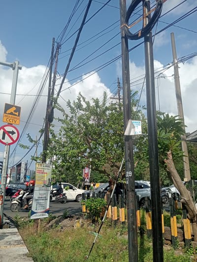 A daytime, eye-level, outdoor medium shot captures a street scene in Surabaya, East Java. The frame is dominated by utility poles and overhead wires, with a bright blue sky and scattered white clouds above. In the foreground, to the left, a sidewalk is visible, bordered by overgrown grass and vegetation. A circular traffic sign with a red border and a black arrow indicating a right turn is prohibited, accompanied by a yellow rectangular sign with a CCTV camera icon and the text "AREA DIAWASI CCTV".

The street level features several vehicles and pedestrians. Two motorcycles, each with a rider wearing a helmet, are traveling on the road. One motorcycle is white and red, the other is black. Multiple cars are also present, including a white SUV and a black sedan. A tall, vertical banner displaying information in Indonesian is attached to a utility pole. To the right of the frame, more utility poles and trees are visible, with a residential building in the background, characterized by its many windows. A black and yellow striped barrier is also present along the side of the road. The overall scene suggests a typical urban environment with ongoing traffic and public services.